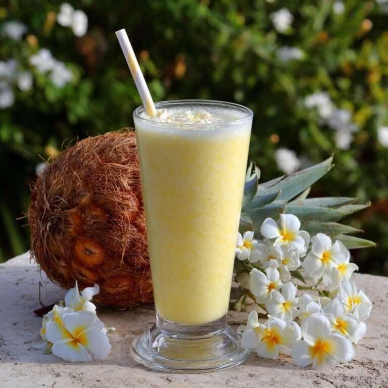 A tall glass with a footed base contains a frothy, light yellow pineapple-coconut smoothie, topped with a white straw and what appears to be shredded coconut. To the left of the glass is a whole pineapple, and behind it, a pineapple crown. In the foreground and to the right, a cluster of white and yellow plumeria flowers are artfully arranged. The setting appears to be outdoors, with a blurred background of green foliage and white flowers under bright, natural light.